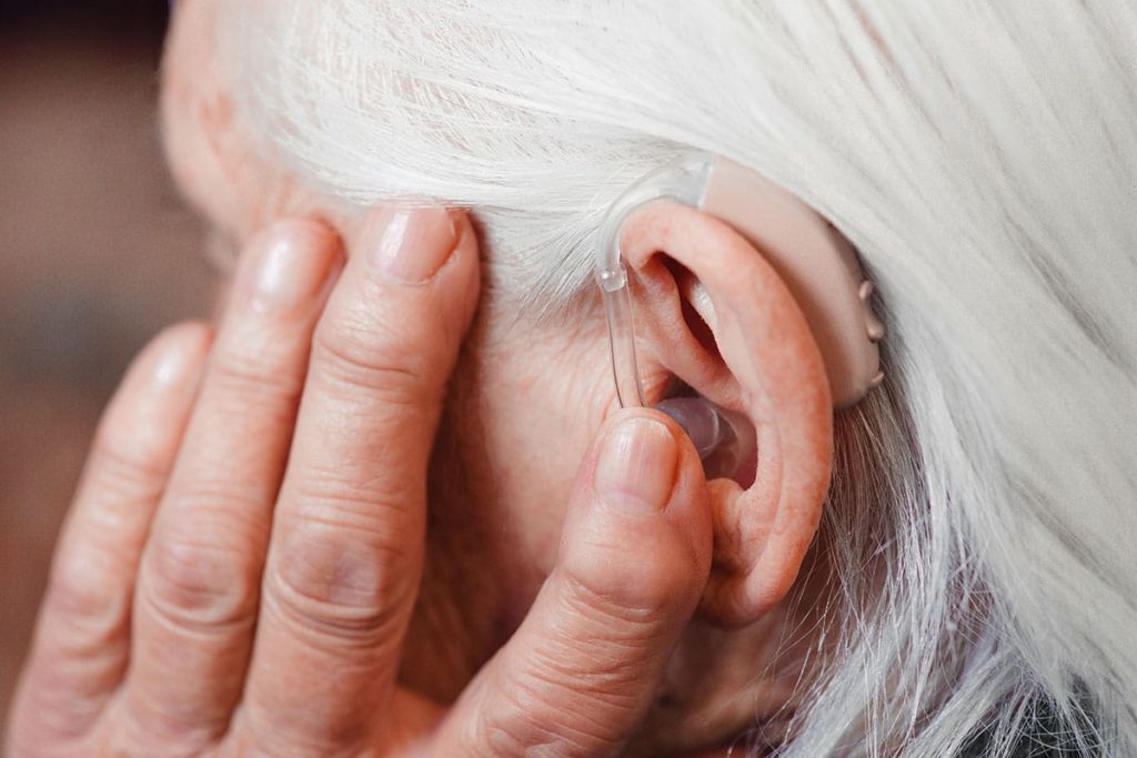 a senior woman wearing a hearing aid touches her hearing aid while wondering the causes of hearing loss