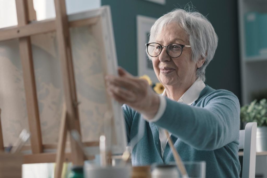 a senior woman is sitting in front of a canvas and painting enjoying one of the many hobbies for seniors
