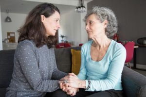 an adult woman holds hands with her senior mother while discussing a memory care community