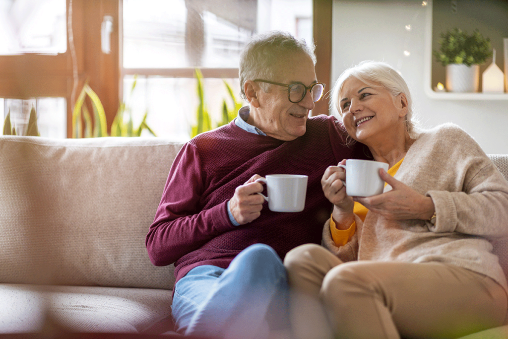 A senior couple is enjoying their coffee together while sitting on a couch in their senior living community.