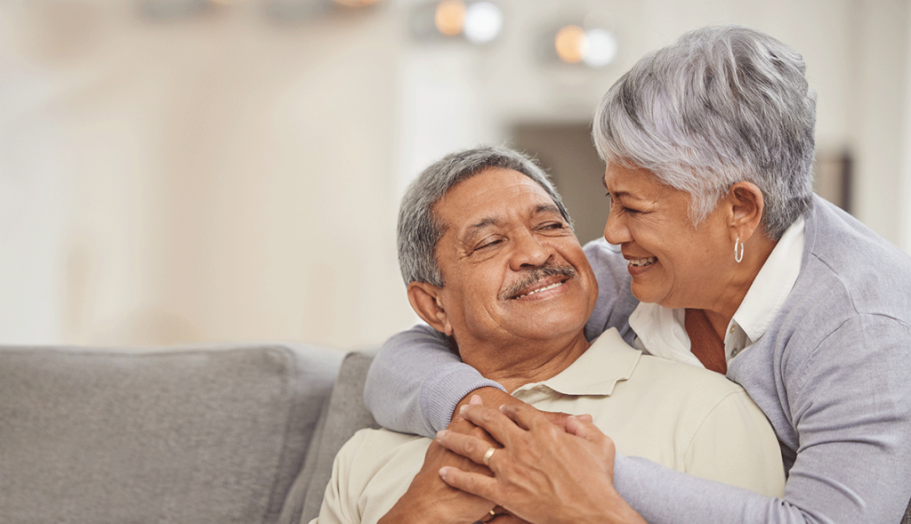 A senior woman is hugging a senior man as he sits on a couch. He's learning how to settle into senior living.