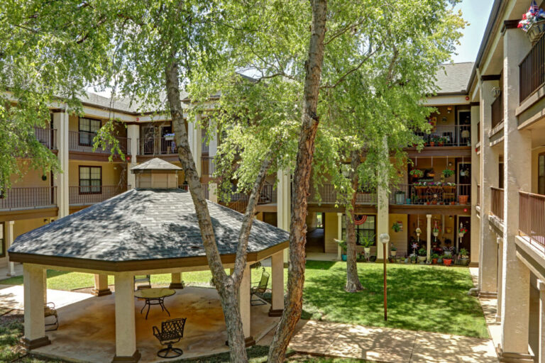 A shaded gazebo with chairs sits in a grassy courtyard, surrounded by a three-story apartment building and tall trees.