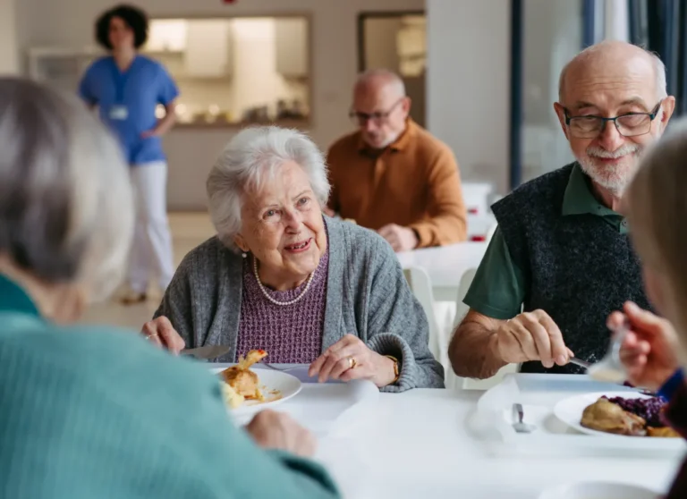 Four seniors eat together in a bright dining area, with a caregiver and another person visible in the background.