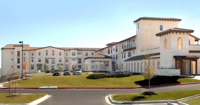 A large, modern, beige building with several windows, cars parked in front, and a well-manicured lawn.