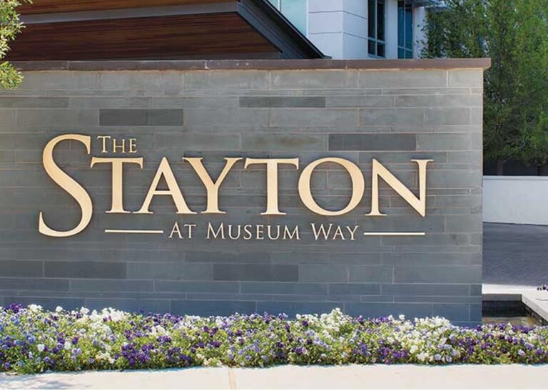 A stone wall sign reads "The Stayton at Museum Way" with flowers planted along the base.