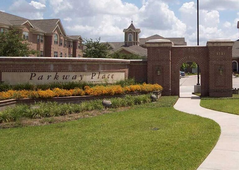 Brick entrance sign reading "Parkway Place" with flowers, pathway, archway, and buildings in the background.