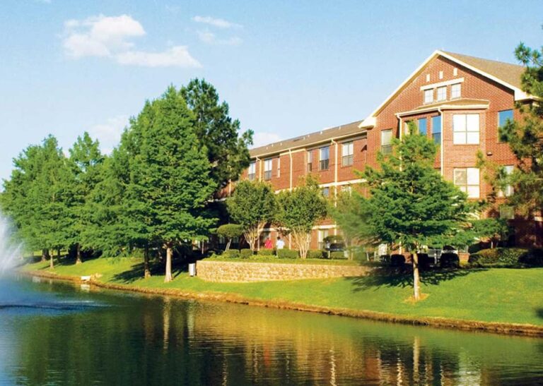 A brick building overlooks a pond with a fountain, surrounded by trees and greenery under a blue sky.