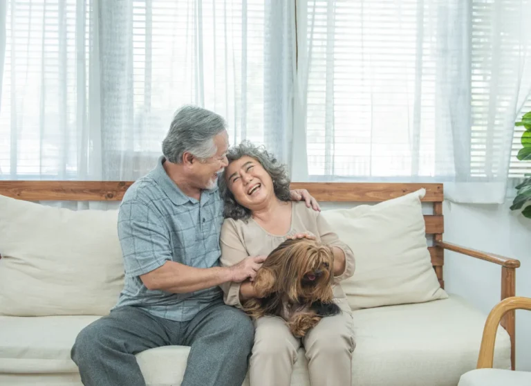 Smiling older couple sits on a couch together, holding and petting a small, long-haired dog in their laps.