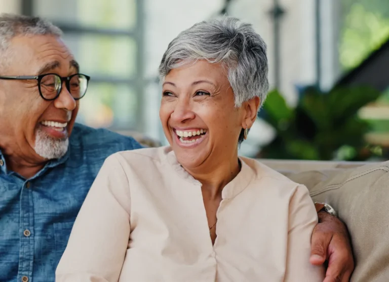 Two older adults sit on a couch, smiling and laughing together in a bright, indoor setting.