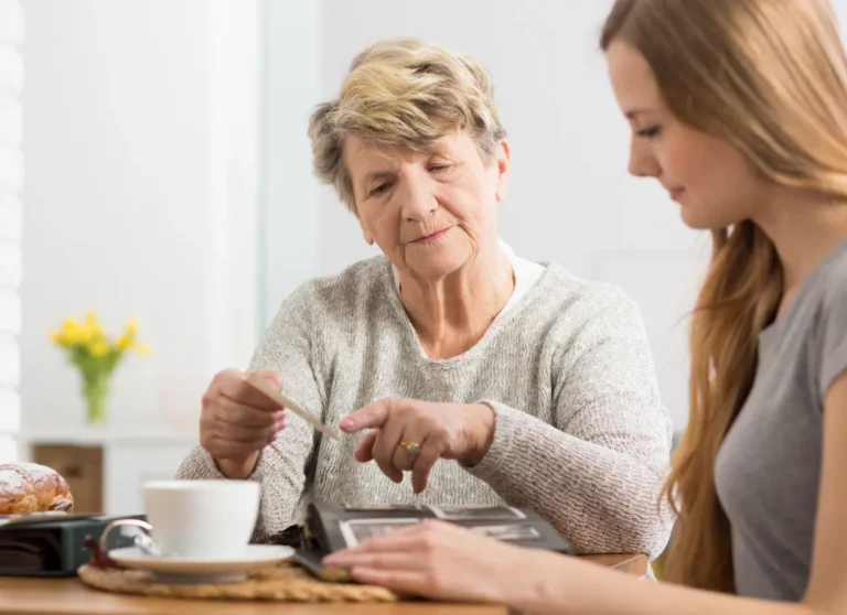 Older woman and younger woman look at a photo album together at a table, sharing a quiet, reflective moment.